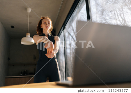 Shooting from below of athletic young woman in outfit doing exercises watching fitness video online on laptop standing by window on sunny day. Female practicing exercise online with laptop 115245377
