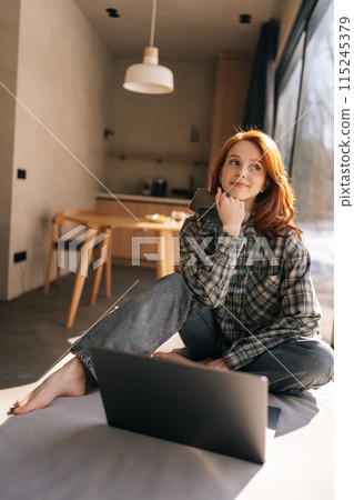 Vertical shot of positive charming young redhead woman dressed in casual clothes relaxing on wide windowsill with portable computer, shopping online, looking away with cute cheerful smile 115245379