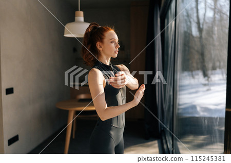 Side view of sporty woman preforming warm-up exercise, stretching arm muscles before workout indoors, standing by window with on sunny day. Athlete prepare muscle for exercise workout at house. 115245381