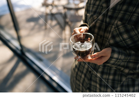 High-angle view of unrecognizable woman holding hot cup of coffee in morning sunlight. Happy satisfied female drinking tea on kitchen. Closeup of lady enjoying cup of coffee at home. High-angle view of unrecognizable woman holding hot cup of coffee in morning sunlight. Happy satisfied female drinking tea on kitchen. Closeup of lady enjoying cup of coffee at home. 115245402