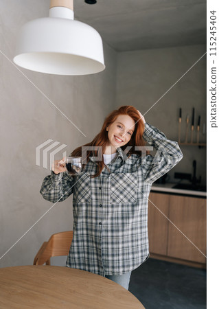 Vertical portrait of cheerful young woman enjoying holding cup drinking coffee standing in kitchen on sunny morning, smiling looking at camera. Happy female relaxing on caffeine break alone. 115245404