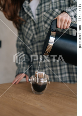 Vertical cropped shot of unrecognizable female pouring hot water in cup with drip coffee bag. Process of making brewed coffee at home. Preparing fresh brewed drip coffee in cup with paper drip bag. 115245415