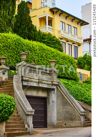 Curving staircase around brown garage door with hedges on roof and yellow building on hill 115245489