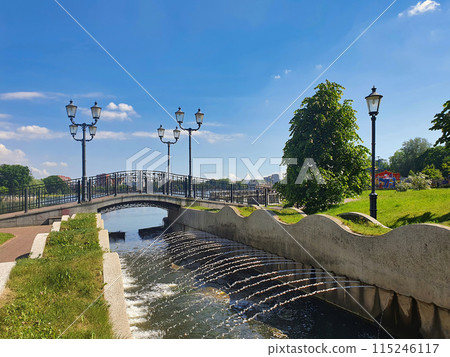 Empty small pedestrian stone bridge over water canal with fountain streams on embankment of Upper Lake - artificial city pond in Kaliningrad, Russia at sunny summer day. With no people cityscape. Empty small pedestrian stone bridge over water canal with fountain streams on embankment of Upper Lake - artificial city pond in Kaliningrad, Russia at sunny summer day. With no people cityscape. 115246117