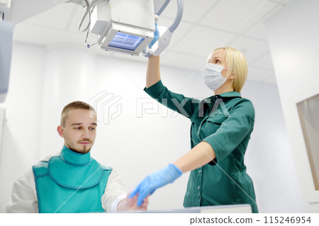 Female radiologist going to made x-ray shot of young man hand in x-ray room in modern clinic. Patient wearing in protect lead apron 115246954