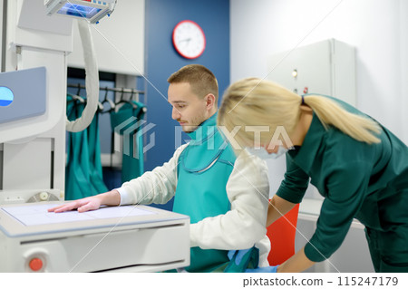 Female radiologist going to made x-ray shot of young man hand in x-ray room in modern clinic. Patient wearing in protect lead apron 115247179