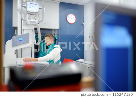Young man having x-ray shot of broken hand in x-ray room in modern clinic. Patient wearing in protect lead apron Young man having x-ray shot of broken hand in x-ray room in modern clinic. Patient wearing in protect lead apron 115247180