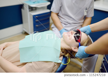 Practitioner dentist doctor prepares woman for teeth treatment in hospital. Assistant wearing protection glasses on patient before orthodontists or prosthetics treatment in modern medical center Practitioner dentist doctor prepares woman for teeth treatment in hospital. Assistant wearing protection glasses on patient before orthodontists or prosthetics treatment in modern medical center 115247210
