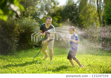 Funny little boy with his father playing with garden hose in sunny backyard. Preschooler child having fun with spray of water. Summer outdoors activity for kids. Funny little boy with his father playing with garden hose in sunny backyard. Preschooler child having fun with spray of water. Summer outdoors activity for kids. 115247339