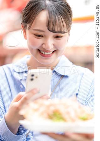 A young, cute, SNS-loving woman taking a photo of takoyaki with her smartphone outdoors in bright light | Dotonbori Shopping Street, Osaka City, Osaka Prefecture 115248588