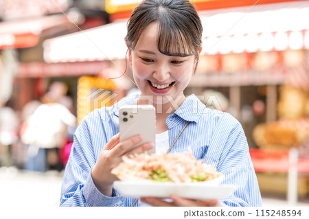 A young, cute, SNS-loving woman taking a photo of takoyaki with her smartphone outdoors in bright light | Dotonbori Shopping Street, Osaka City, Osaka Prefecture 115248594