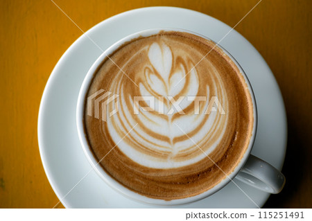 A detailed close-up of latte with intricate leaf art in white cup, set on yellow wooden table 115251491