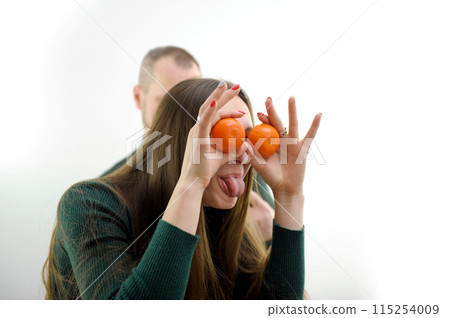 young girl put Tangerines to eyes and pulled out tongue stuck out and she twists and makes faces in the background a man on a white background a green sweater long hair joke fooling around grimacing young girl put Tangerines to eyes and pulled out tongue stuck out and she twists and makes faces in the background a man on a white background a green sweater long hair joke fooling around grimacing 115254009