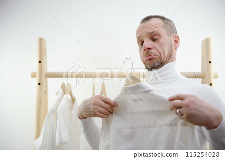 man against background of wooden hanger grimaces trying on putting shirt on himself doesnot like even frowned eyebrows closed eyes on hand ring concept of choosing clothes tailoring cleaning laundry 115254028