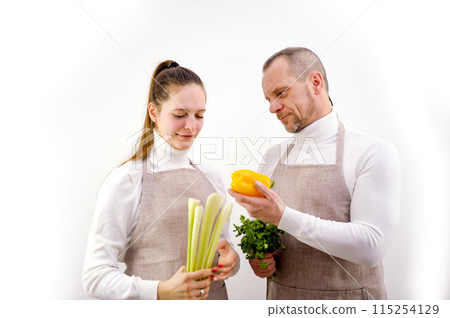 husband wife in kitchen aprons look at vegetables people's clothes and background are white they have peppers their hands celery parsley cilantro they are vegetarians fresh vegetables cook dinner husband wife in kitchen aprons look at vegetables people's clothes and background are white they have peppers their hands celery parsley cilantro they are vegetarians fresh vegetables cook dinner 115254129