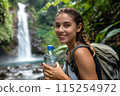 happy young woman tourist with backpack holding bottle of water against the backdrop of mountain waterfall 115254972