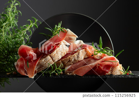 Ciabatta with prosciutto and rosemary on a black background. Ciabatta with prosciutto and rosemary on a black background. 115256925