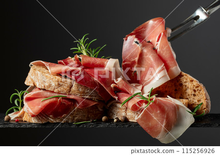 Ciabatta with prosciutto and rosemary on a black background. Ciabatta with prosciutto and rosemary on a black background. 115256926