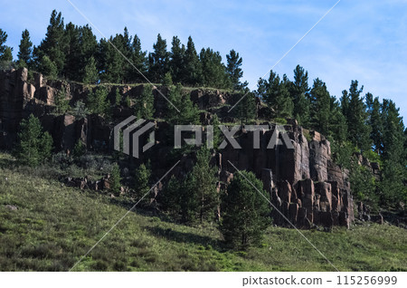 Blue sky with white clouds over the mountains. The rocks in the form of sandstone pillars are overgrown with pine trees. Mountain summer landscape. Wildlife of Eastern Siberia. Blue sky with white clouds over the mountains. The rocks in the form of sandstone pillars are overgrown with pine trees. Mountain summer landscape. Wildlife of Eastern Siberia. 115256999