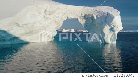 Arch in melting Arctic iceberg floating blue ocean water. Low angle drone flight in big hole white snow covered ice glacier. Ecology, conservation, melting ice, climate change, global warming concept 115259343