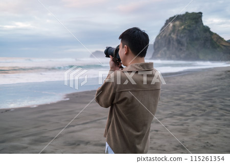 Male tourist is taking a photo with Lion Rock on Piha Beach in the evening at West of Auckland, New Zealand 115261354