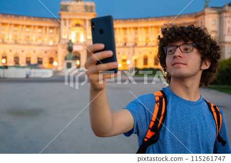 Vienna, Austria, August 17, 2022. A young Caucasian man with curly hair and glasses takes a selfie in front of the Hofburg Imperial Palace at sunset. The warm evening light enhances the imposing Vienna, Austria, August 17, 2022. A young Caucasian man with curly hair and glasses takes a selfie in front of the Hofburg Imperial Palace at sunset. The warm evening light enhances the imposing 115261747
