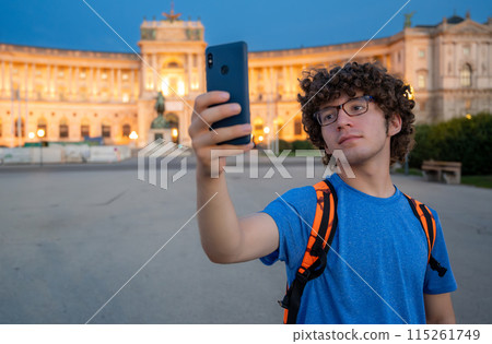 Vienna, Austria, August 17, 2022. A young Caucasian man with curly hair and glasses takes a selfie in front of the Hofburg Imperial Palace at sunset. The warm evening light enhances the imposing 115261749
