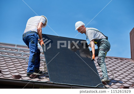 Workers building solar panel system on roof of house. Two men installers in helmets carrying photovoltaic solar module outdoors. Alternative, green and renewable energy generation concept. Workers building solar panel system on roof of house. Two men installers in helmets carrying photovoltaic solar module outdoors. Alternative, green and renewable energy generation concept. 115261929
