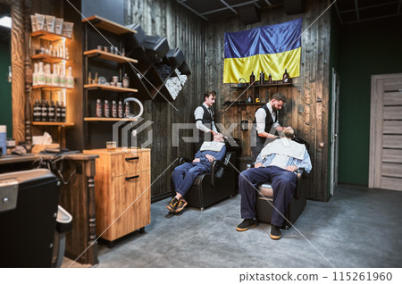 Professional barbers washing hair with deep cleaning shampoo. Busy day in modern Ukrainian barbershop. Male clients relaxing while getting their hair done. 115261960