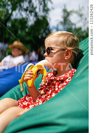 Little girl eating a banana. Portrait of a fashionable, stylish beautiful baby girl in sunglasses and a red dress while sitting in a bean bag chair. Little girl eating a banana. Portrait of a fashionable, stylish beautiful baby girl in sunglasses and a red dress while sitting in a bean bag chair. 115262426