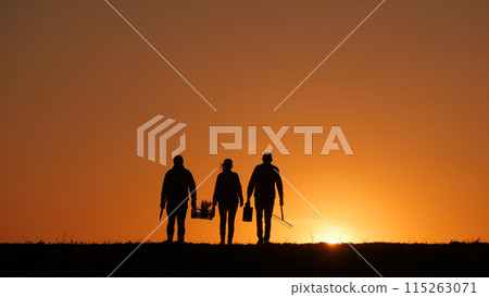 Three farmers with equipment walk through a field at sunset 115263071