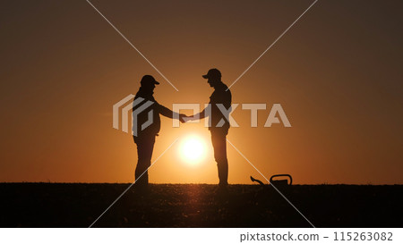 Two farmers' silhouettes shake hands with the sunset over the field as the backdrop. 115263082