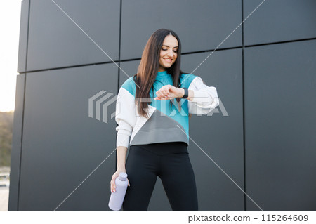 Sporty young woman checks results of fitness training and holds bottle with fresh water, dressed in activewear poses outdoors. Healthy lifestyle Sporty young woman checks results of fitness training and holds bottle with fresh water, dressed in activewear poses outdoors. Healthy lifestyle 115264609
