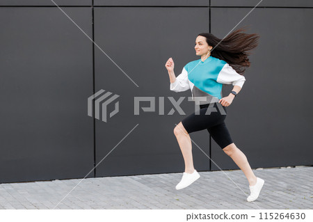 Young woman with fit body jumping and running against dark wall background. Female model in sportswear exercising outdoors. Young woman with fit body jumping and running against dark wall background. Female model in sportswear exercising outdoors. 115264630