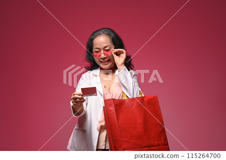 Portrait happy mature woman holding shopping bag and credit card over red background 115264700