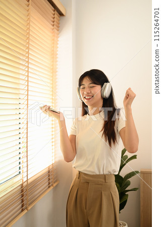 Happy young asian woman holding phone listening to favorite songs and dancing in living room 115264701