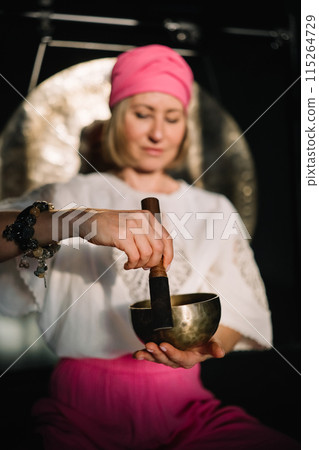 Middle aged woman using singing bowl in sound healing therapy indoors.  115264729