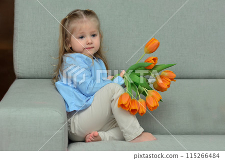 A three-year-old girl sits on the sofa and holds orange tulips in her hands. A three-year-old girl sits on the sofa and holds orange tulips in her hands. 115266484