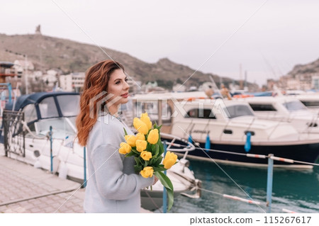 Woman holds yellow tulips in harbor with boats docked in the background., overcast day, yellow sweater, mountains 115267617
