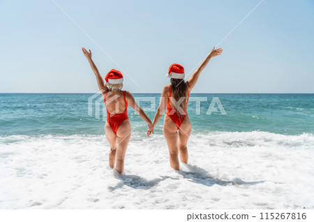 Women Santa hats ocean play. Seaside, beach daytime, enjoying beach fun. Two women in red swimsuits and Santa hats are enjoying themselves in the ocean waves and raising their hands up. Women Santa hats ocean play. Seaside, beach daytime, enjoying beach fun. Two women in red swimsuits and Santa hats are enjoying themselves in the ocean waves and raising their hands up. 115267816