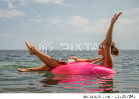 A woman is floating in a pink inflatable raft with a laptop in her hand. She is smiling and she is enjoying her time in the water. 115267846