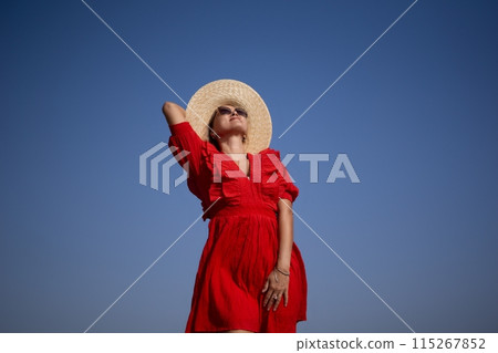 A woman in a red dress is wearing a straw hat and standing in the sun 115267852