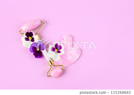 Pink jade roller, gua sha for face massage with purple and white flower viola. Facial massager tools. Anti age, lifting and toning care. Modern selfcare concept. Pink jade roller, gua sha for face massage with purple and white flower viola. Facial massager tools. Anti age, lifting and toning care. Modern selfcare concept. 115268683