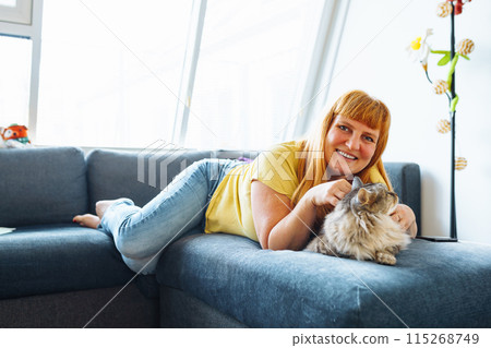 portrait positive blonde middle-aged woman lying on sofa hugging cat taking selfie 115268749