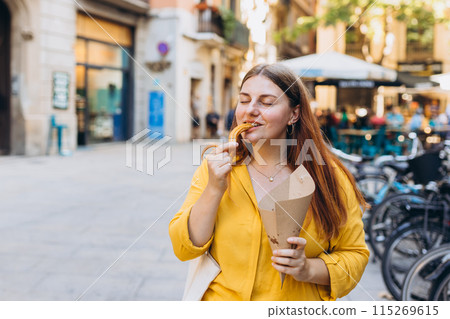 Carefree woman eating traditional Churros, a fried pastry with chocolate on a city street. Positive cheerful female student posing outdoors. Food concept 115269615