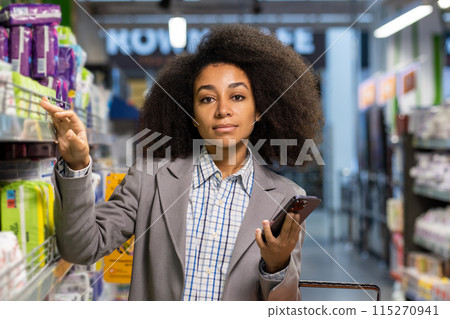Confident woman in a supermarket aisle holding a smartphone and reaching for an item on the shelf. Confident woman in a supermarket aisle holding a smartphone and reaching for an item on the shelf. 115270941