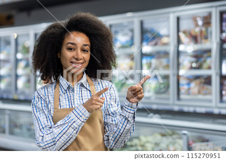 A cheerful supermarket employee in an apron, smiling and pointing at products in the frozen food aisle. Retail and customer service concept. A cheerful supermarket employee in an apron, smiling and pointing at products in the frozen food aisle. Retail and customer service concept. 115270951