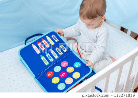 Little toddler girl playing with busy book while sitting in crib. Concept of quiet books and modern educational toys for kids Little toddler girl playing with busy book while sitting in crib. Concept of quiet books and modern educational toys for kids 115271839