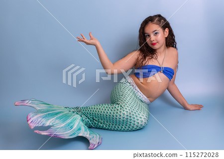 A beautiful elegant girl in a mermaid costume lies on a blue background with her palm raised up. A beautiful elegant girl in a mermaid costume lies on a blue background with her palm raised up. 115272028