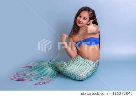 A happy girl in a mermaid costume sitting on a blue background showing thumbs up. 115272045